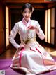 A woman in a white and pink hanbok sitting on a purple mat.